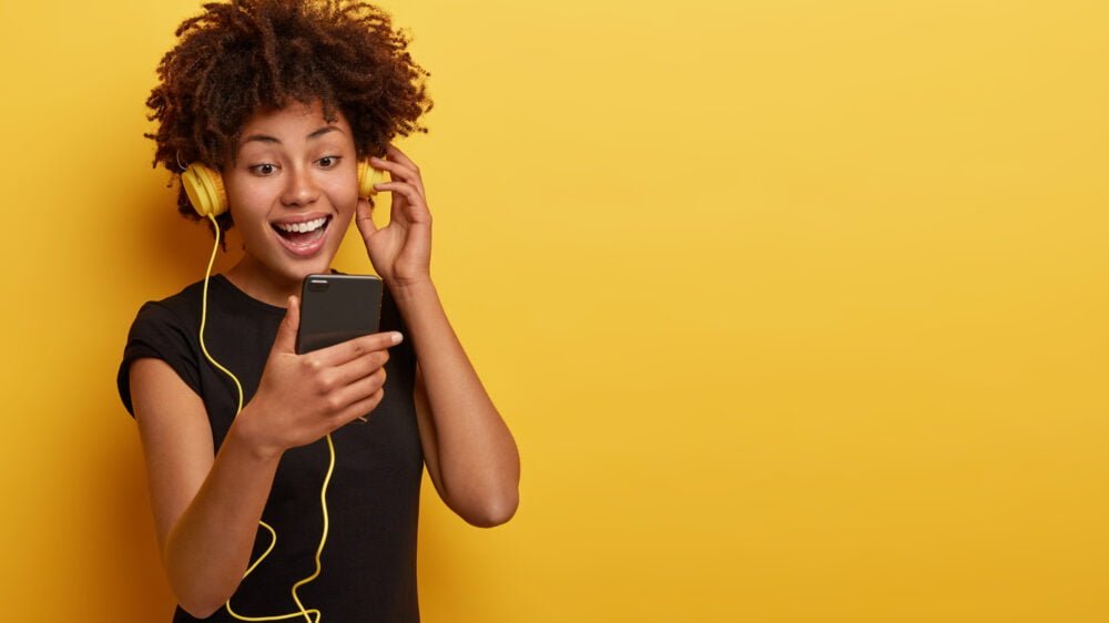 happy girl focused in cell phone, enjoys listening music, happy to renew playlist, uses special app, smiles broadly, dressed in black t shirt, stands indoor over yellow wall empty space, watches video